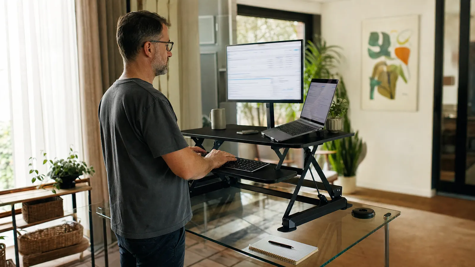 Home office desk with a black two-tier standing desk converter raised to full height, keyboard tray extended, adult typing while standing at a glass-top desk