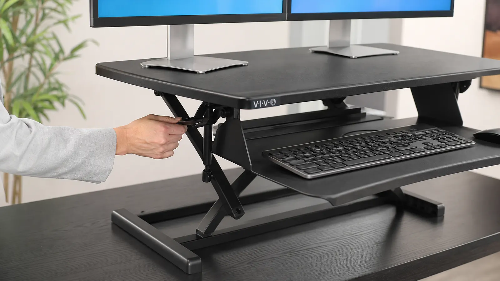 Close-up of a hand raising a black dual-level desk converter riser on top of a wood desk, laptop and monitor resting on the upper platform