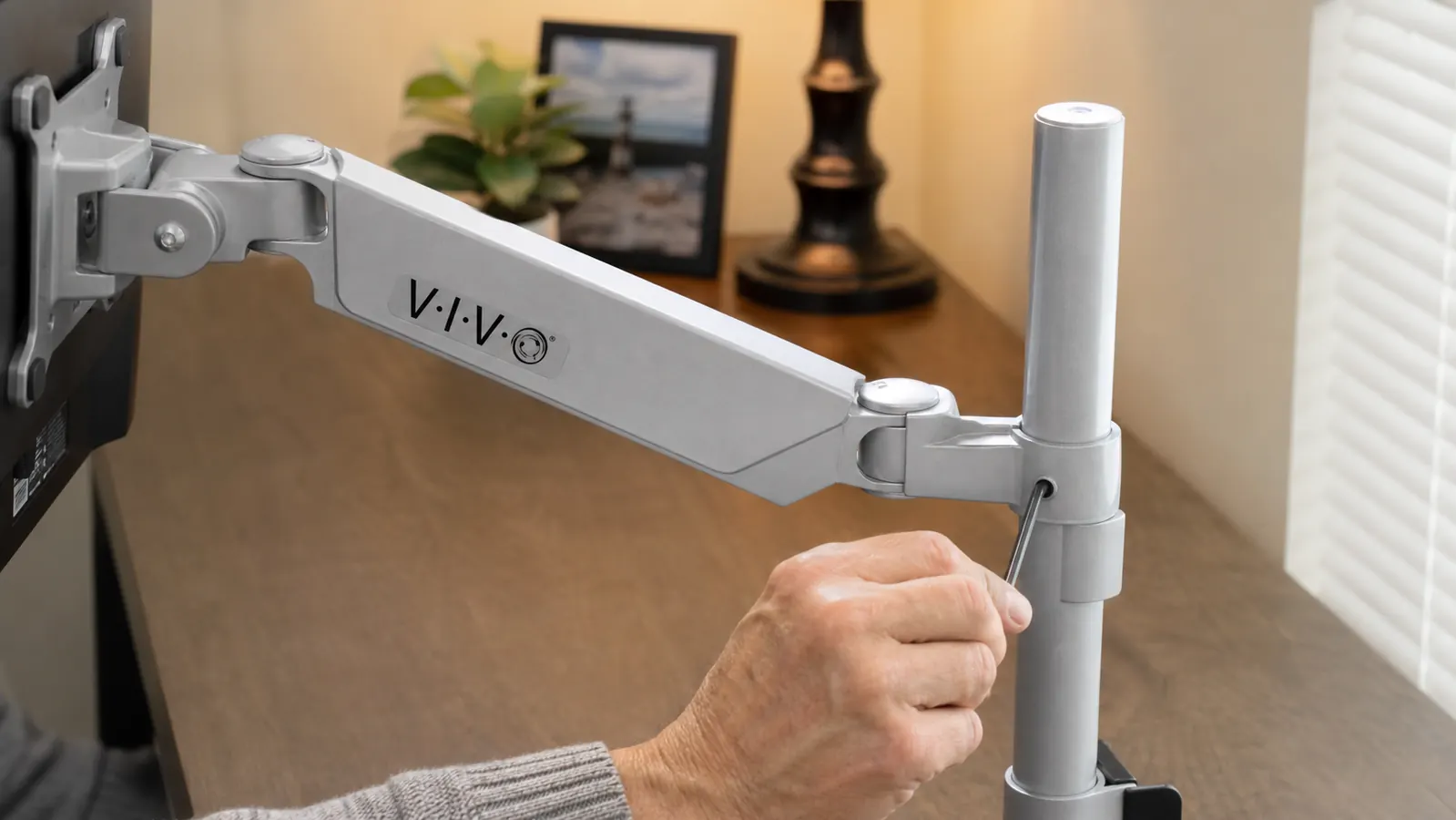 Hand turning a hex key inside the vertical joint of a monitor arm to adjust spring tension, with the arm extended out over a desk